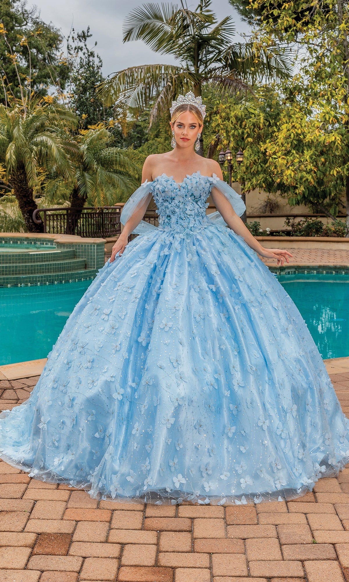 A woman in the Quinceanera Dress 1820 By Dancing Queen, featuring a blue ball gown with floral details and a sweetheart neckline, wears a tiara as she stands by a swimming pool surrounded by trees and greenery.